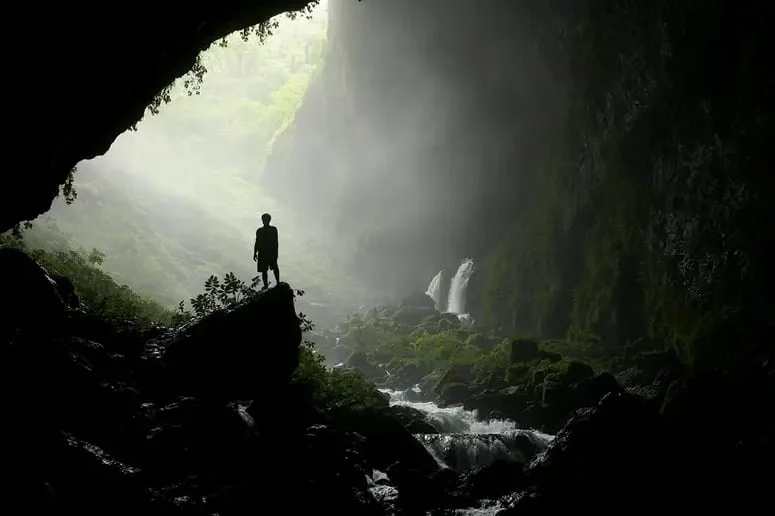 A person standing at the mouth of a vast cave with a waterfall
