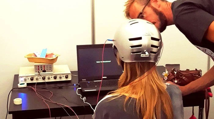 A researcher fitting a helmet on a participant who sits at a laptop